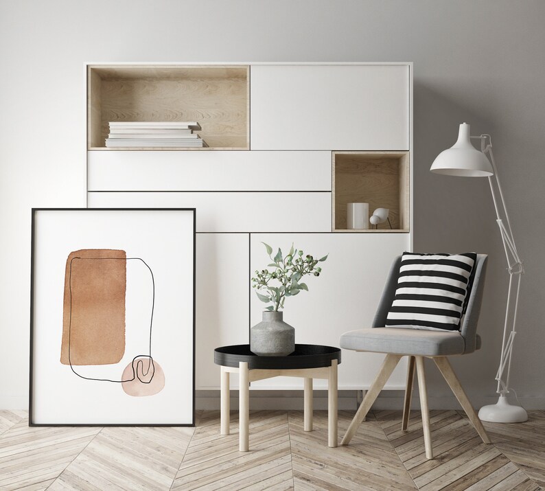 Op de afbeelding: Een witte kast met een zwart ingelijst abstract kunstwerk, een kleine ronde houten tafel met een zwarte dienblad, een vaas met groene bladeren en een grijze stoel met een zwart-wit gestreept kussen. De kast heeft een plank met boeken en een klein open vak met een witte vaas en een klein wit voorwerp.