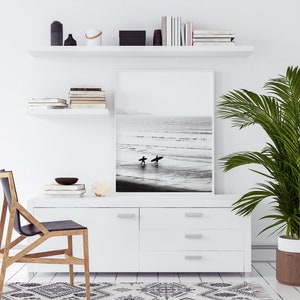 May include: A black and white photograph of two surfers walking on a beach with their surfboards. The photo is framed and hanging on a white wall above a white dresser with three drawers. A potted plant is on the right side of the image.