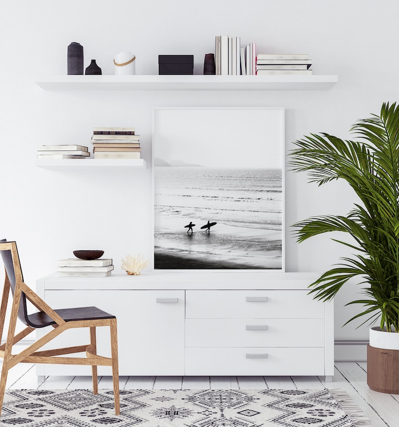 May include: A black and white photograph of two surfers walking on a beach with their surfboards. The photo is framed and hanging on a white wall above a white dresser with three drawers. A potted plant is on the right side of the image.