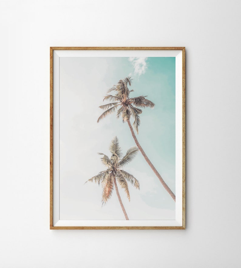 May include: A framed print of two palm trees against a light blue sky. The trees are silhouetted against the sky, with their fronds reaching up towards the top of the frame.