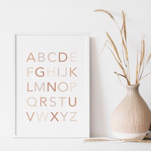 May include: A white framed print with the alphabet in a gradient of beige and brown colors. The letters are arranged in rows, with the first row being "ABCDE", the second row being "FGHIJK", and so on. The print is hanging on a white wall, and there is a vase of dried grass in the foreground.