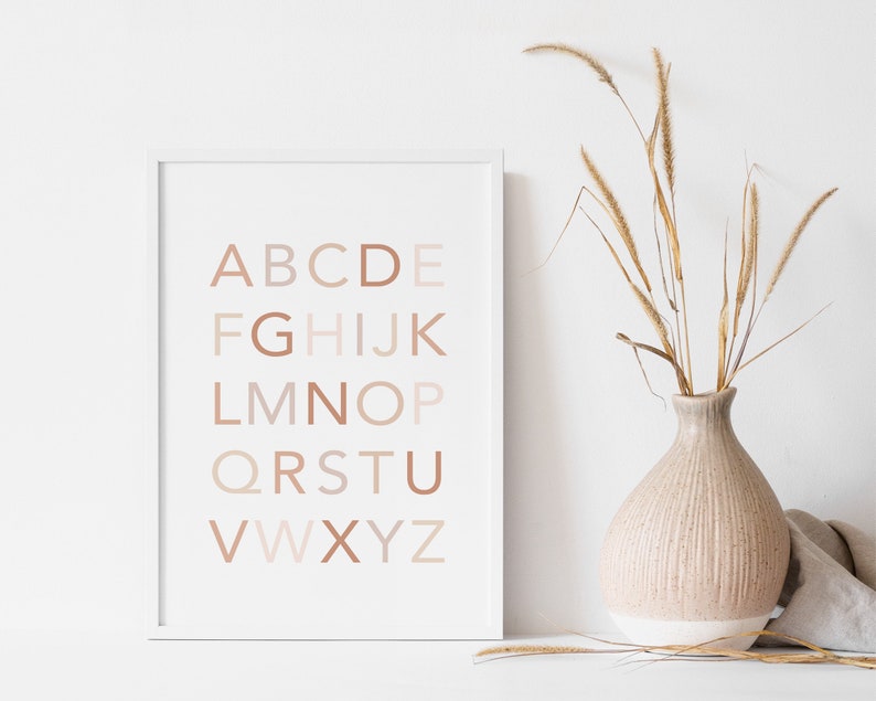 May include: A white framed print with the alphabet in a gradient of beige and brown colors. The letters are arranged in rows, with the first row being "ABCDE", the second row being "FGHIJK", and so on. The print is hanging on a white wall, and there is a vase of dried grass in the foreground.