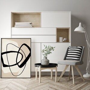 May include: A black and white abstract art print with a beige background hangs on a white wall in a living room setting. A small side table with a black top and wooden legs sits in front of the print. A grey chair with a black and white striped cushion sits next to the table. A white floor lamp with a white shade stands in the corner of the room.