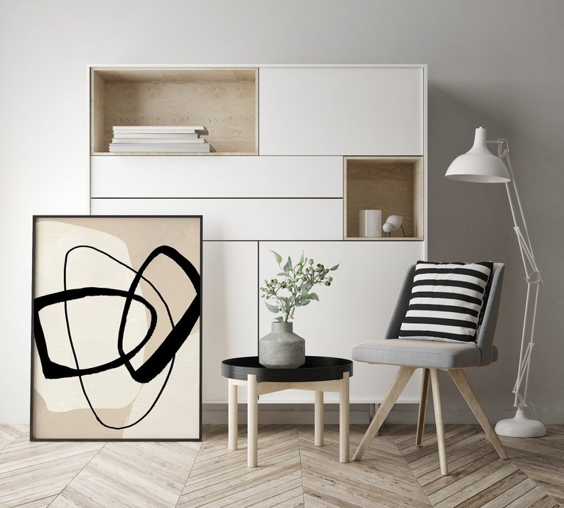 May include: A black and white abstract art print with a beige background hangs on a white wall in a living room setting. A small side table with a black top and wooden legs sits in front of the print. A grey chair with a black and white striped cushion sits next to the table. A white floor lamp with a white shade stands in the corner of the room.