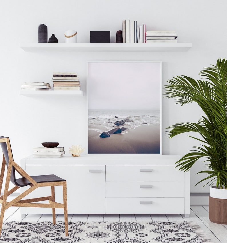 May include: A white dresser with four drawers and silver handles. A framed photograph of a beach scene with rocks and waves hangs above the dresser. A potted palm plant is on the right side of the image. A black and white patterned rug is on the floor in front of the dresser.