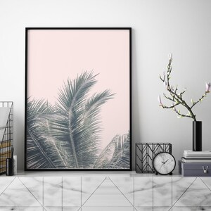 May include: A black framed print of a palm tree with green leaves against a light pink background. The print is on a white marble shelf with a black vase with a white flower branch, a black metal magazine rack, and a black clock.