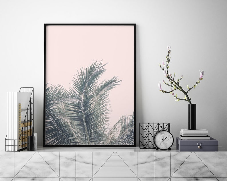 May include: A black framed print of a palm tree with green leaves against a light pink background. The print is on a white marble shelf with a black vase with a white flower branch, a black metal magazine rack, and a black clock.