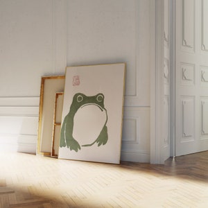 May include: A framed print of a green frog on a white background. The print is leaning against a white wall and a wooden floor.