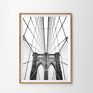 Pu&ograve; includere: Fotografia in bianco e nero del ponte di Brooklyn, incorniciata in una cornice di legno marrone chiaro. L'intricata rete di cavi e torri del ponte &egrave; visibile contro un cielo bianco brillante. L'immagine &egrave; una composizione verticale.