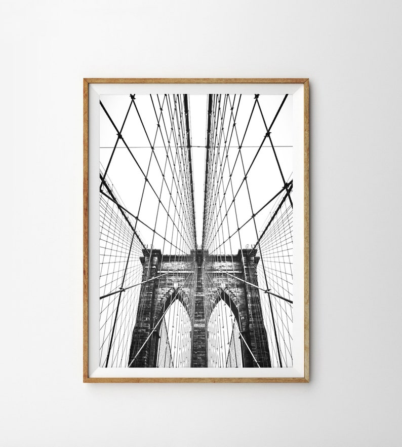 Pu&ograve; includere: Fotografia in bianco e nero del ponte di Brooklyn, incorniciata in una cornice di legno marrone chiaro. L'intricata rete di cavi e torri del ponte &egrave; visibile contro un cielo bianco brillante. L'immagine &egrave; una composizione verticale.