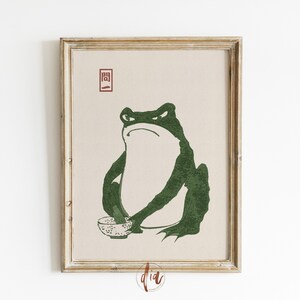 May include: A framed print of a green frog holding a bowl. The frog is sitting on a beige background with the Japanese character "開" in red.