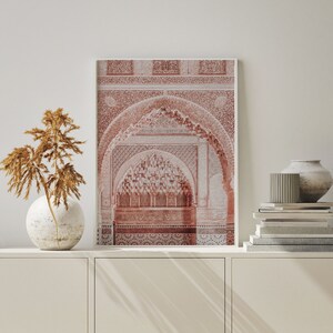 May include: A framed print of a red and white intricate archway with a detailed pattern. The archway is set against a white wall with a vase of dried flowers and a stack of books on a white shelf.
