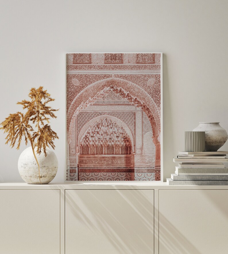 May include: A framed print of a red and white intricate archway with a detailed pattern. The archway is set against a white wall with a vase of dried flowers and a stack of books on a white shelf.