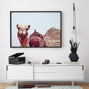 May include: A framed photograph of a camel with a colorful saddle in front of a sand dune. The photo is hanging on a white wall above a white dresser with a record player, headphones, and books on top.