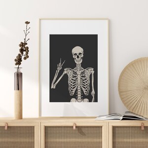 May include: A framed print of a skeleton with a skull making a peace sign. The skeleton is white and the background is black.