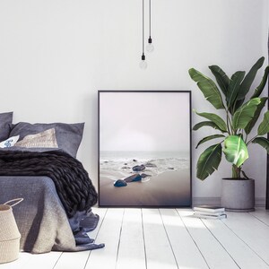 May include: A bedroom interior with a gray bed, a black knit throw blanket, and a framed photograph of a beach scene with rocks and waves. A potted plant with large green leaves is on the right side of the image.