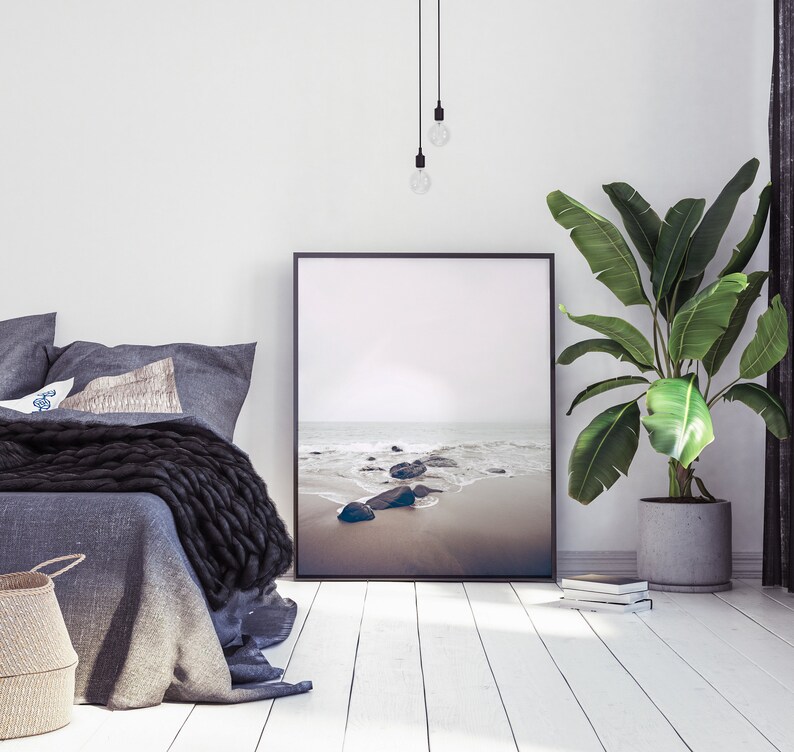 May include: A bedroom interior with a gray bed, a black knit throw blanket, and a framed photograph of a beach scene with rocks and waves. A potted plant with large green leaves is on the right side of the image.