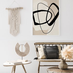 May include: A beige and black abstract art print with a white frame hangs above a wooden bench with a black and white cushion. A wooden beaded necklace sits on a small wooden table next to a book. A vase with dried pampas grass sits on a woven wicker table.
