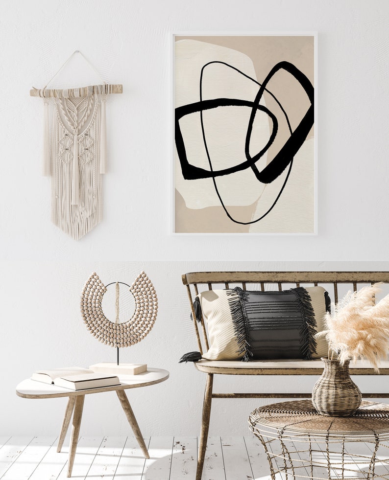May include: A beige and black abstract art print with a white frame hangs above a wooden bench with a black and white cushion. A wooden beaded necklace sits on a small wooden table next to a book. A vase with dried pampas grass sits on a woven wicker table.