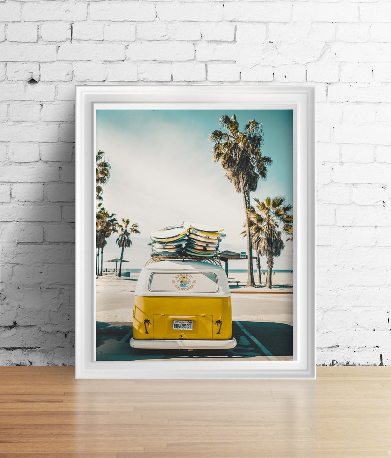 May include: A yellow vintage van with surfboards on the roof parked in front of a beach with palm trees. The van has a white stripe and the words "KAUAI SURF CLUB" on the back.