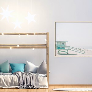 May include: A wooden bunk bed with a gray blanket and blue pillows. The bed is decorated with string lights and white star-shaped decorations. A framed print of a beach scene with a lifeguard stand is on the wall.
