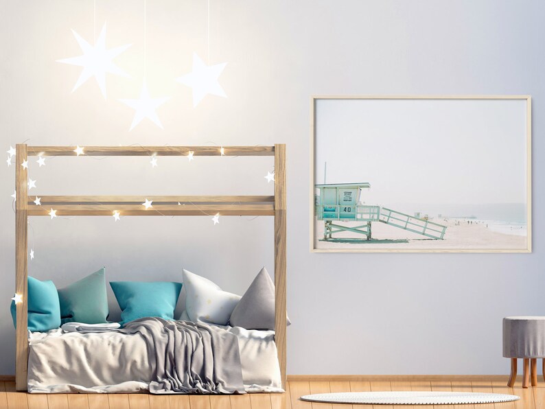 May include: A wooden bunk bed with a gray blanket and blue pillows. The bed is decorated with string lights and white star-shaped decorations. A framed print of a beach scene with a lifeguard stand is on the wall.