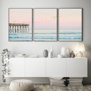 May include: Three framed prints depicting a beach scene with a pier in the distance and a flock of birds flying overhead. The sky is a soft pink and blue, and the water is a turquoise blue.
