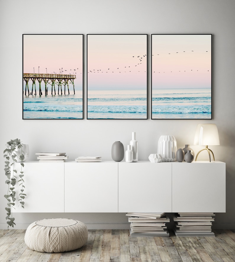 May include: Three framed prints depicting a beach scene with a pier in the distance and a flock of birds flying overhead. The sky is a soft pink and blue, and the water is a turquoise blue.