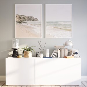May include: Two framed beach landscape prints hang above a white cabinet. The cabinet is styled with a black candle holder, a potted plant, decorative vases, books, and a desk lamp. The prints depict a sandy beach and ocean.