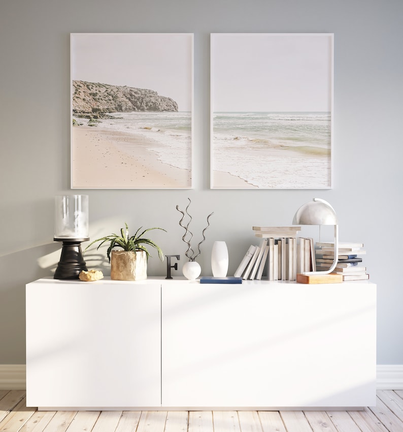 May include: Two framed beach landscape prints hang above a white cabinet. The cabinet is styled with a black candle holder, a potted plant, decorative vases, books, and a desk lamp. The prints depict a sandy beach and ocean.