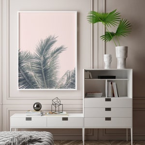 May include: A white framed print of a palm tree against a pink background hangs on a wall above a white dresser with drawers and shelves. The dresser is next to a white console table with a black geometric sculpture and a black orb on top.