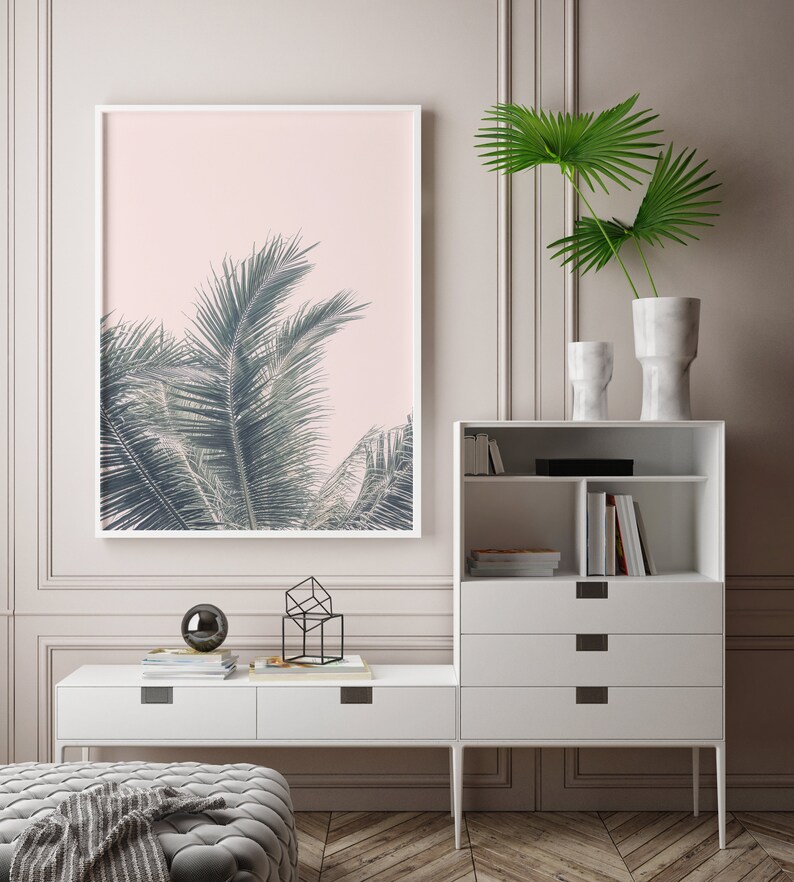 May include: A white framed print of a palm tree against a pink background hangs on a wall above a white dresser with drawers and shelves. The dresser is next to a white console table with a black geometric sculpture and a black orb on top.