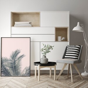 May include: A white shelf unit with a light wood interior, featuring a framed print of a palm tree against a pink background, a small potted plant, and a gray chair with a black and white striped cushion. The chair is positioned in front of a small wooden table.