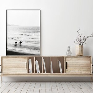 May include: A wooden media console with two sliding doors, open shelves, and black hardware. A black framed photograph of two surfers walking on a beach is displayed above the console. A vase with a small flowering branch sits on top of the console.
