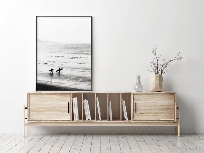May include: A wooden media console with two sliding doors, open shelves, and black hardware. A black framed photograph of two surfers walking on a beach is displayed above the console. A vase with a small flowering branch sits on top of the console.