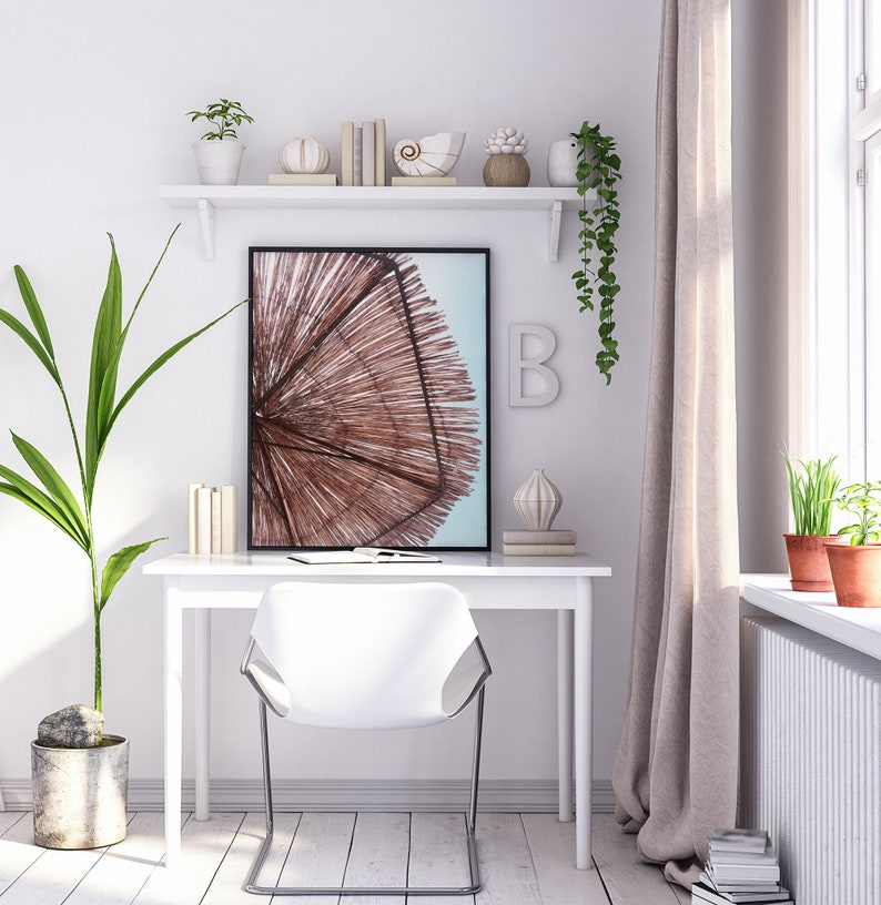 May include: A white desk with a white chair and a framed print of a beach umbrella. The desk has a shelf above it with decorative items, including books, a seashell, and a plant. There is a plant on the floor next to the desk and a window with curtains in the background.