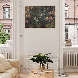 May include: A framed floral painting featuring pink and white roses, displayed on a white wall in a living room. The room includes a beige sofa, a wooden coffee table, and a potted plant. Windows on either side offer views of buildings and greenery.
