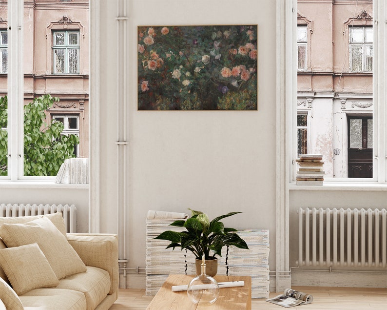 May include: A framed floral painting featuring pink and white roses, displayed on a white wall in a living room. The room includes a beige sofa, a wooden coffee table, and a potted plant. Windows on either side offer views of buildings and greenery.