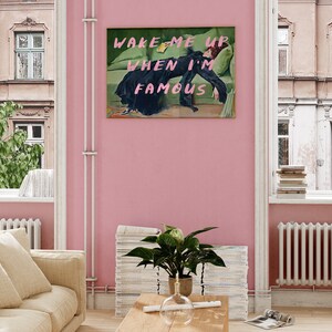 May include: A pink wall with a framed print of a woman lying on a green couch. The print says "WAKE ME UP WHEN I'M FAMOUS".
