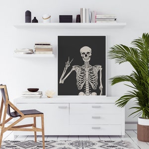 May include: A black and white illustration of a skeleton giving the peace sign, printed on a black background. The illustration is framed and hanging on a white wall above a white dresser.