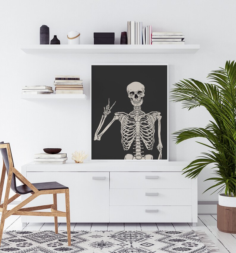 May include: A black and white illustration of a skeleton giving the peace sign, printed on a black background. The illustration is framed and hanging on a white wall above a white dresser.