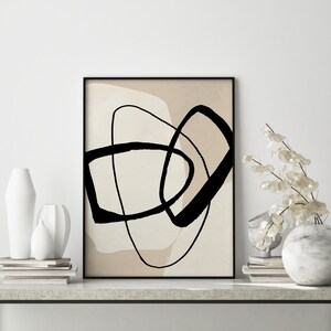 May include: Abstract art print featuring black lines and shapes against a beige background. The print is framed in black and is displayed on a white wall.