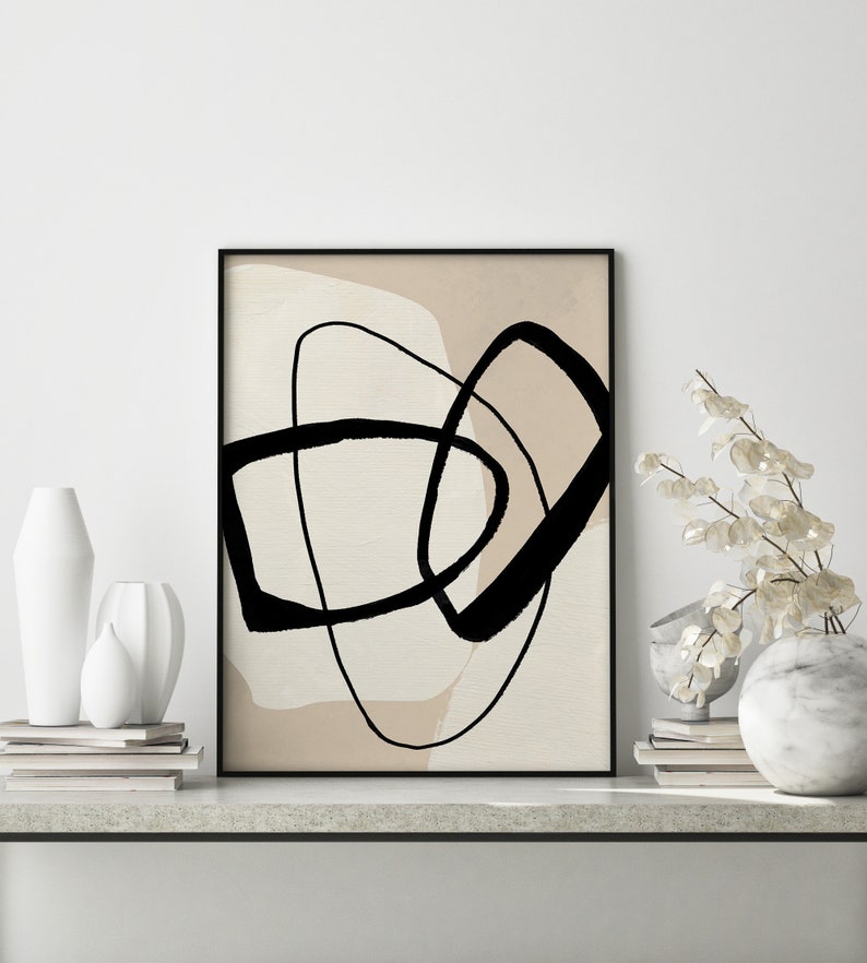 May include: Abstract art print featuring black lines and shapes against a beige background. The print is framed in black and is displayed on a white wall.