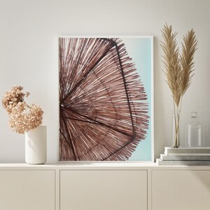 May include: A framed print of a brown woven beach umbrella against a blue sky. The umbrella is made of natural materials and has a rustic look.