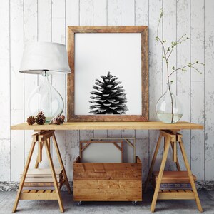 May include: A black and white photograph of a pine cone in a wooden frame, hanging on a white wall above a wooden table with two wooden trestles. The table is decorated with a lamp, two small pine cones, and a vase with a branch.