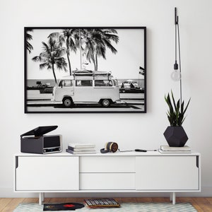 May include: Black and white photo of a vintage van parked on a beach with palm trees in the background. The photo is framed in black and hanging on a white wall above a white cabinet with a record player and headphones on top.