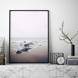 May include: A black framed photograph of a beach scene with rocks and waves. The photo is hanging on a white wall above a marble shelf with a black vase with white flowers, a black metal magazine holder, and a white clock.