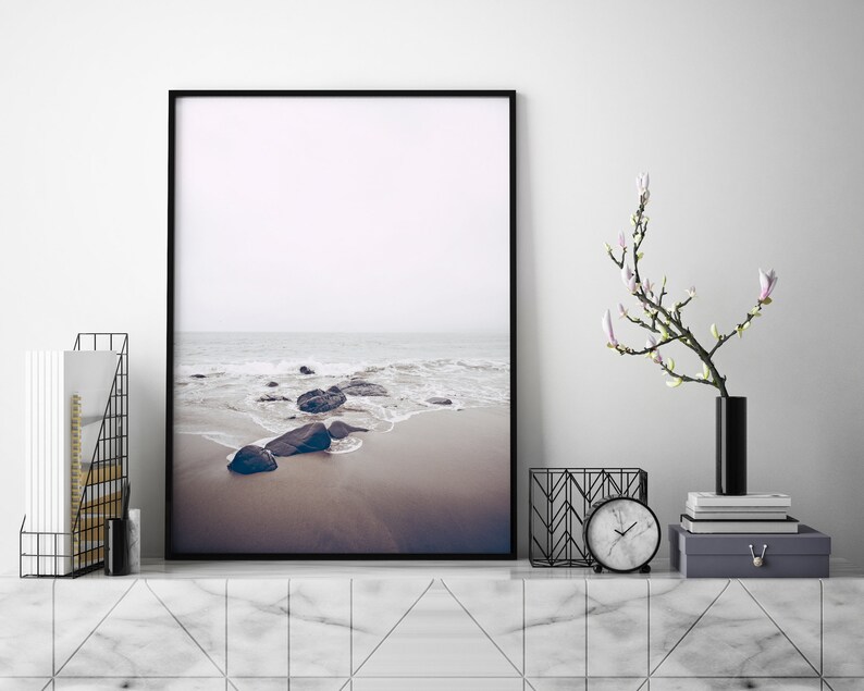 May include: A black framed photograph of a beach scene with rocks and waves. The photo is hanging on a white wall above a marble shelf with a black vase with white flowers, a black metal magazine holder, and a white clock.