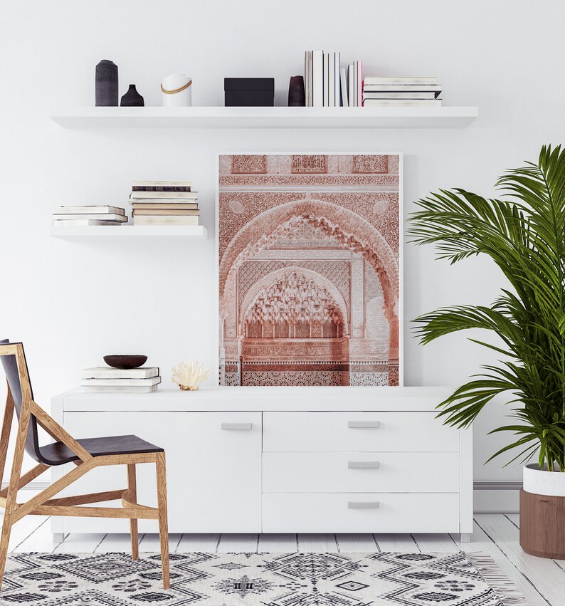 May include: A white shelf with books and decorative objects, a white dresser with drawers, a black chair, a potted plant, and a framed print of a red archway with intricate details.