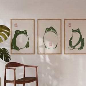 May include: Three framed prints of green frogs on a white background. The frogs are in different poses, one is sitting, one is standing, and one is holding a bowl. The prints are in wooden frames.